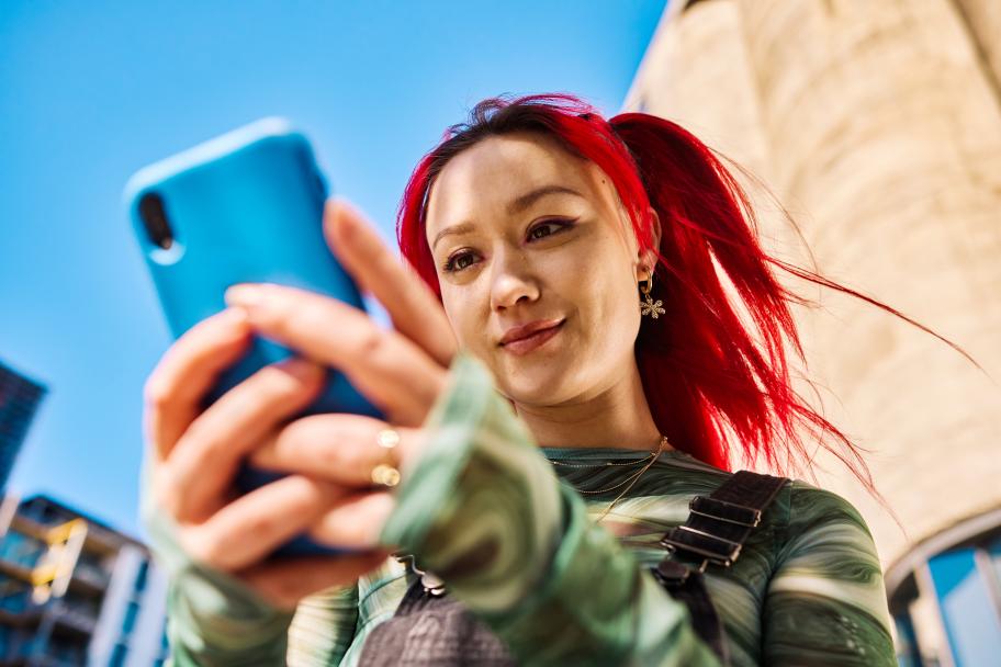 A person with dyed red hair wearing a green jacket and looking at a cell phone.